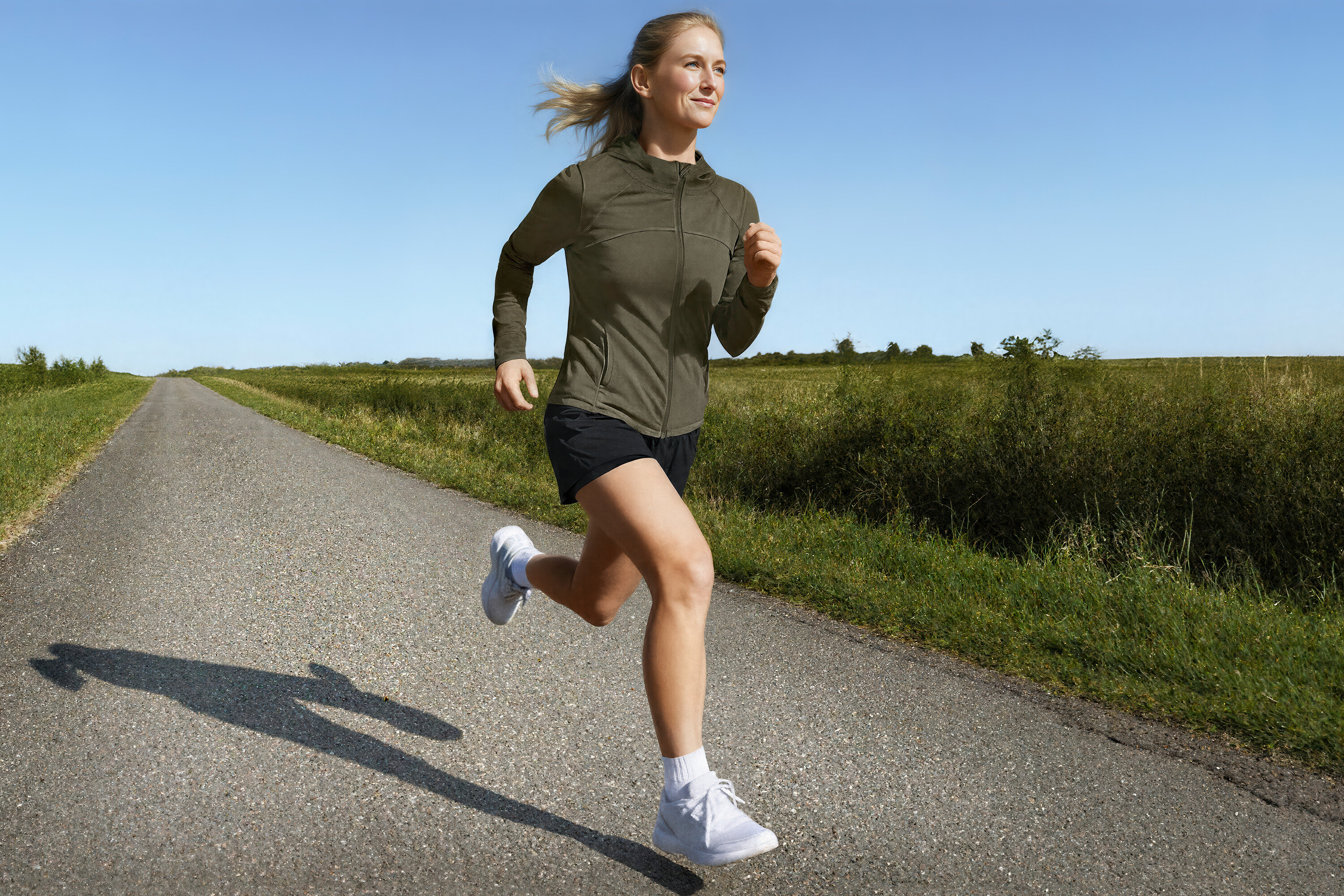 Frau läuft in Sportjacke und 2-in-1-Laufshorts auf einer Landstraße.