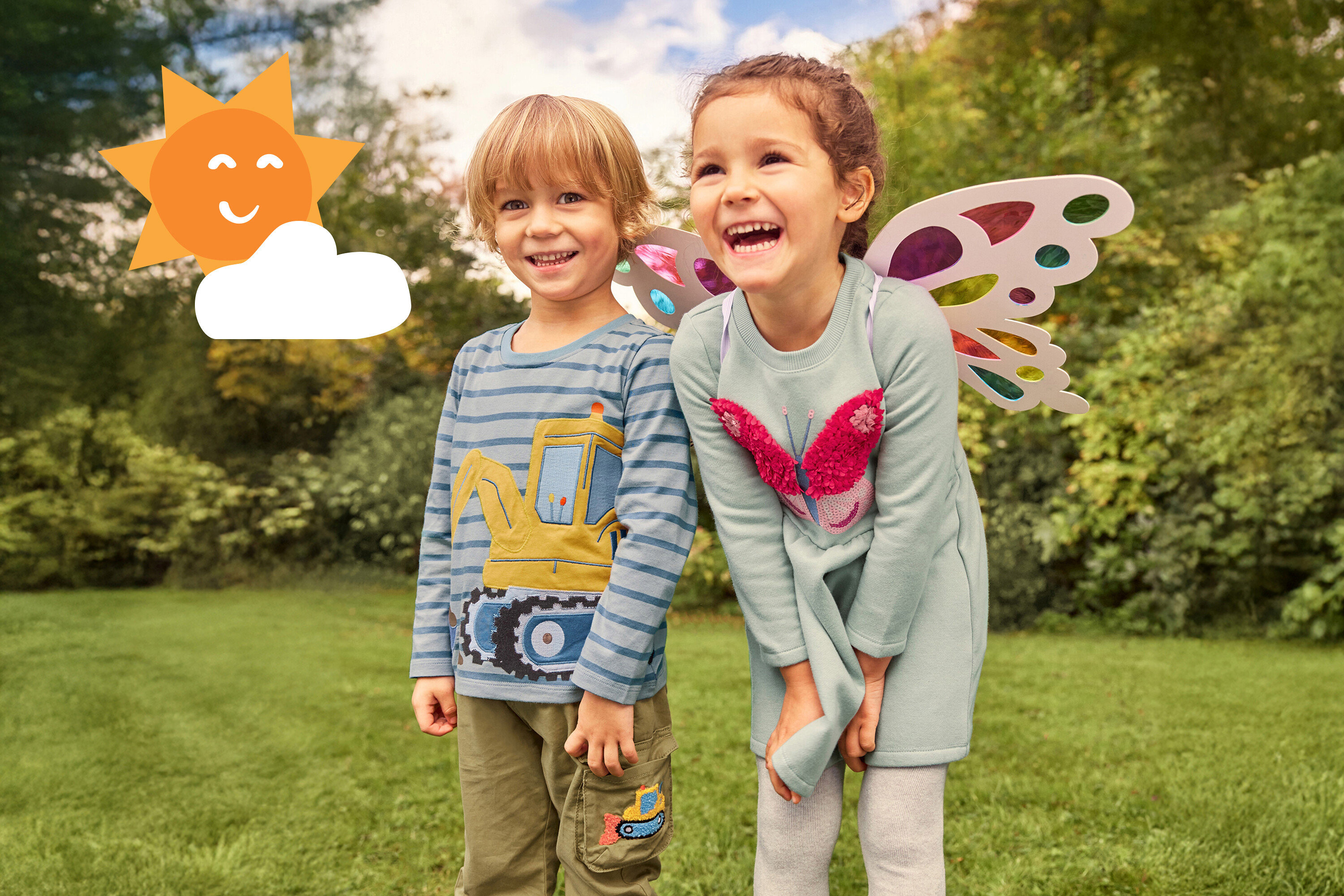 Zwei lachende Kinder stehen im Gras. Das Mädchen trägt Flügel und ein Kleid mit Schmetterling, der Junge ein Bagger-Shirt.
