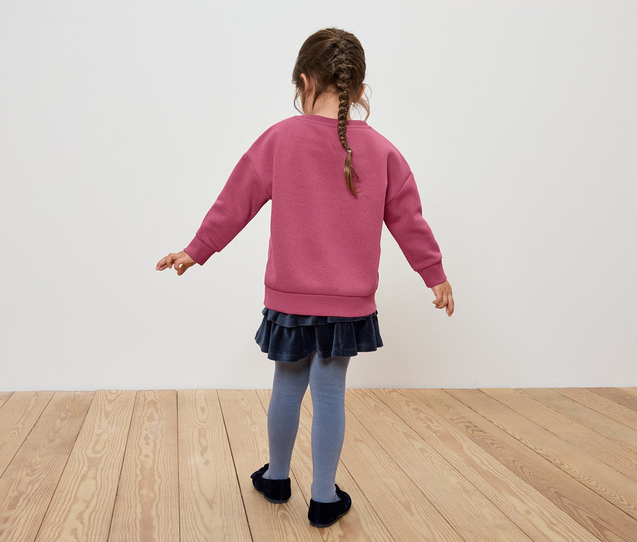 Mädchen mit Zopf steht mit dem Rücken zur Kamera und trägt ein rosa Sweatshirt, einen blauen Rock und graue Strumpfhosen.