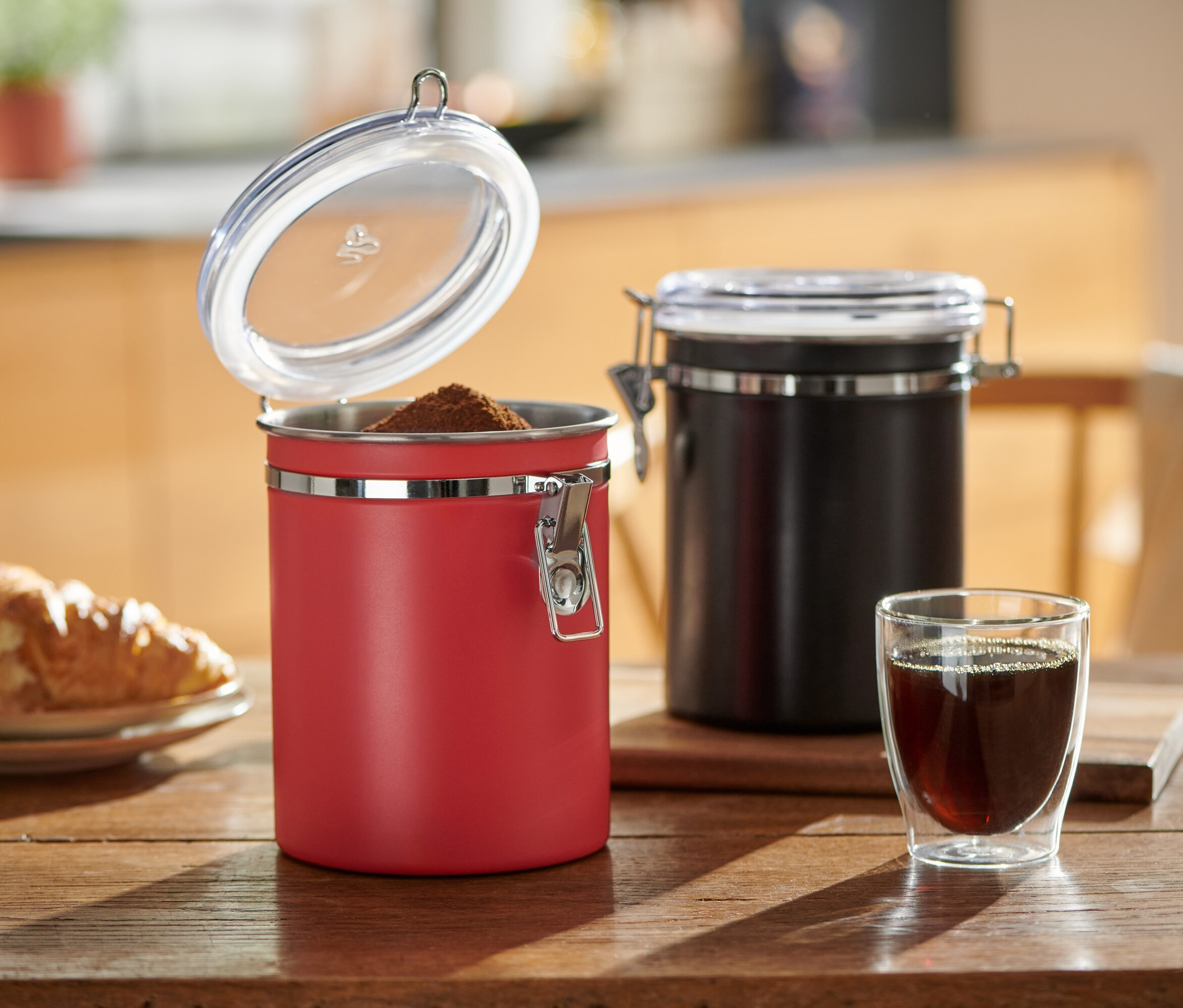 Auf einem Holztisch stehen eine offene rote und eine geschlossene schwarze Aromadose neben einem Glas Kaffee und einem Teller mit einem Croissant.