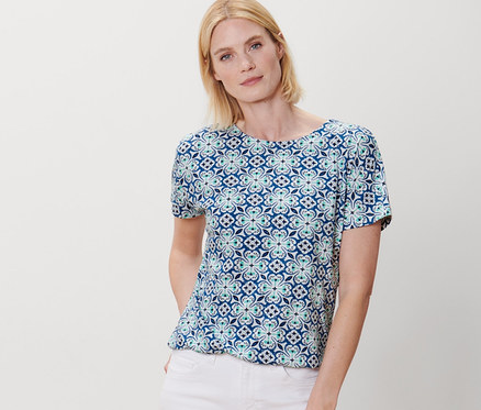 Frau posiert in einem Blusenshirt mit Alloverprint und weissen Hosen.