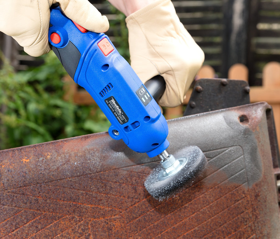 Eine Hand mit Handschuh benutzt eine blaue Westfalia Akku-Poliermaschine auf einer verrosteten Metalloberfläche.