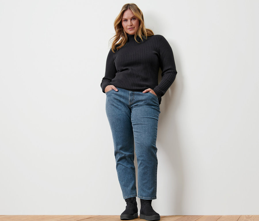 Eine Frau steht mit den Händen in den Hosentaschen ihrer Jeans. Sie trägt einen gerippten Rollkragenpullover, schwarz.