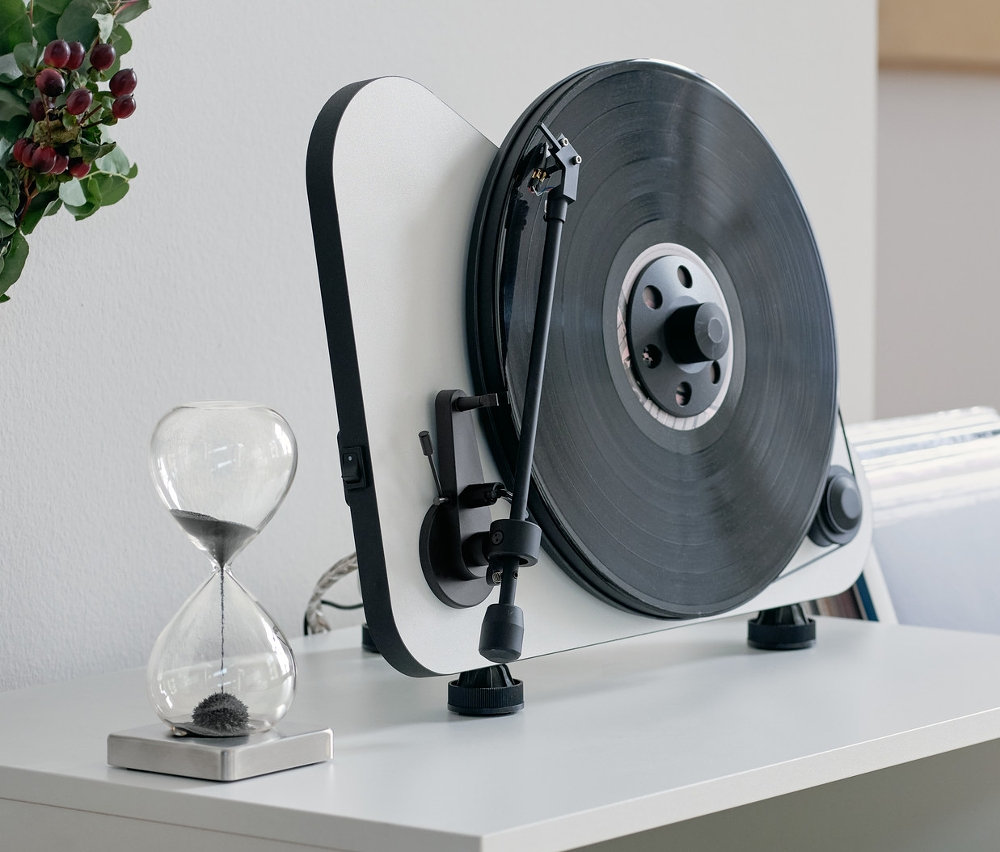 Auf einer weißen Kommode stehen eine magnetische Sanduhr, ein Stereo Regallautsprecher mit Bluetooth® und ein Vertikaler Plug-and-Play-Plattenspieler »Pro-Ject VT-E BT« mit Bluetooth®.