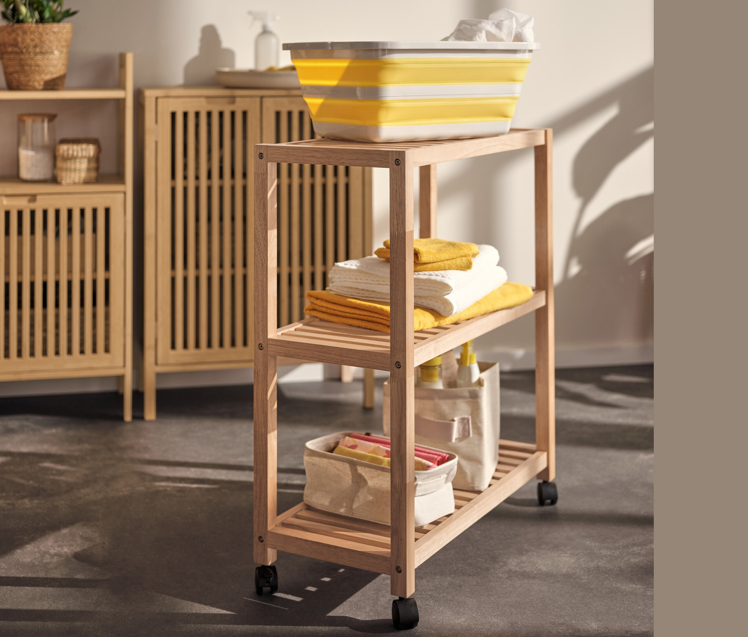 Gelber Wäschekorb auf einem Massivholzregal mit Rollen neben einem Sideboard.