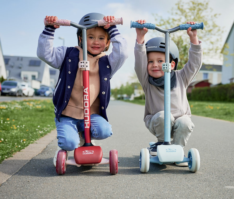 Zwei Kinder knien auf der Straße auf HUDORA Tri-Scootern, einer bordeauxrot, der andere blau.