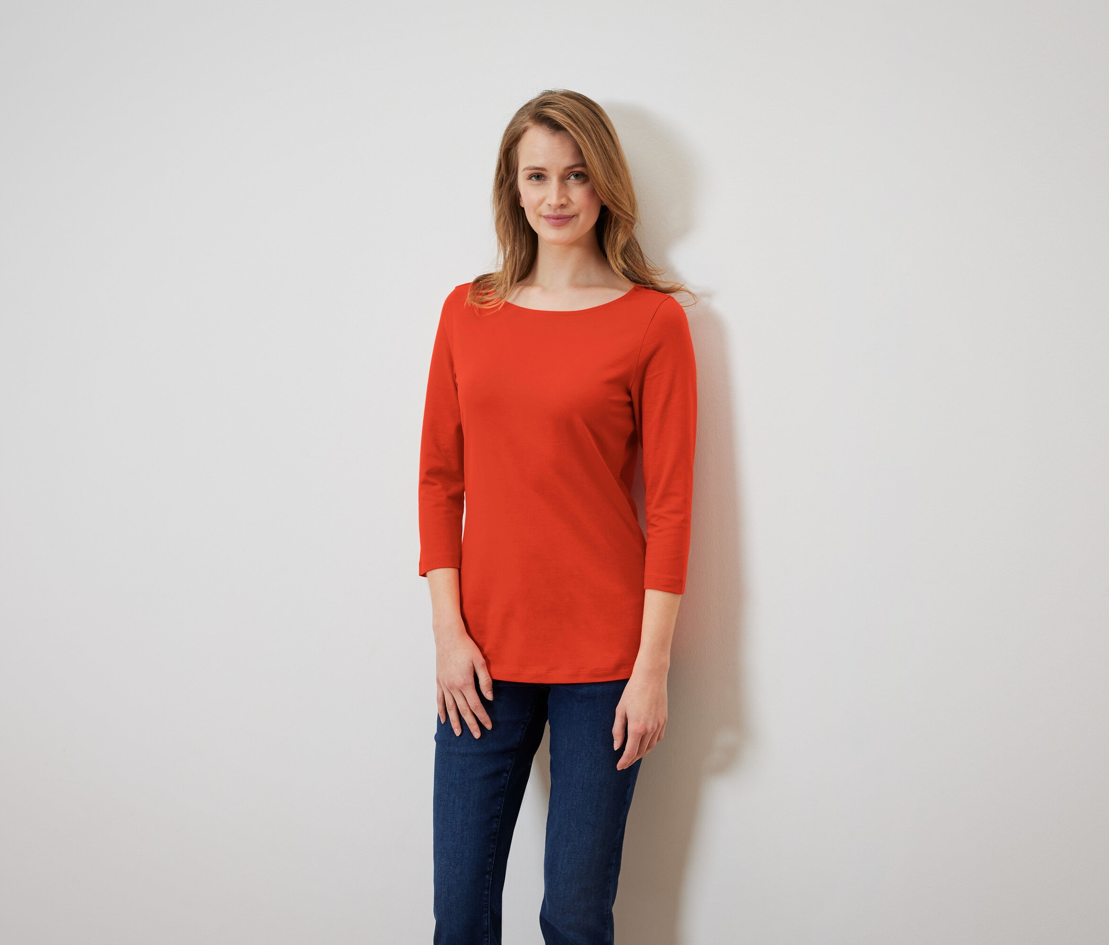 Frau mit langen Haaren steht und trägt ein orangefarbenes Dreiviertelarm-Shirt und Jeans.
