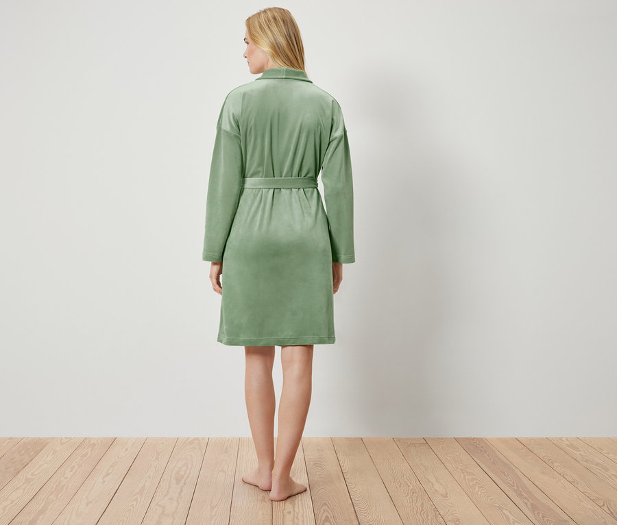 Rückansicht einer blonden Frau in einem hellgrünen Bademantel, die auf einem Holzboden vor einer weißen Wand steht.