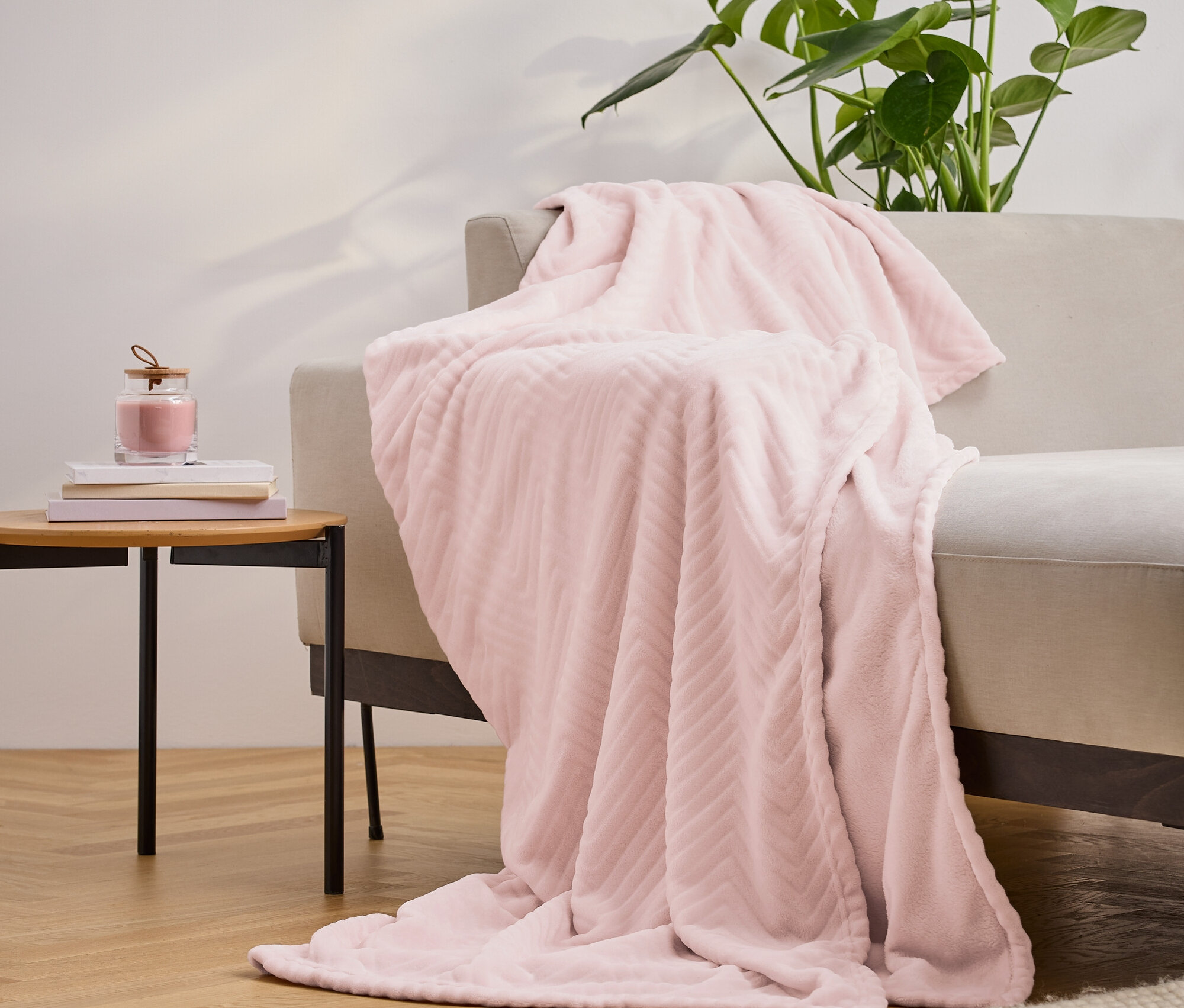 Eine rosa, strukturierte Decke liegt über einem beige farbenen Sofa. Daneben steht ein Tisch mit Büchern und einer Kerze, im Hintergrund eine Pflanze.