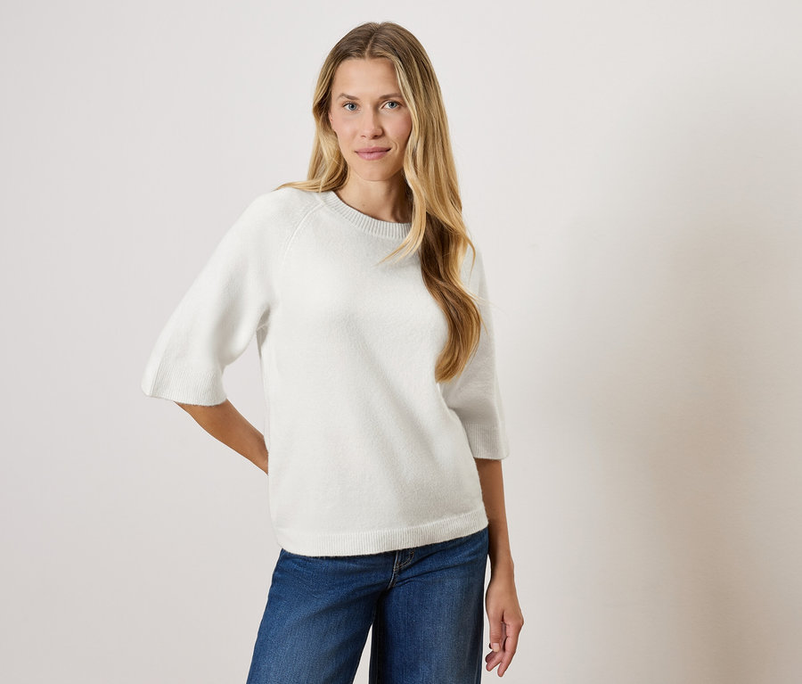 Frau mit langen blonden Haaren posiert in einem weißen Pullover mit Dreiviertelärmeln und blauen Jeans.