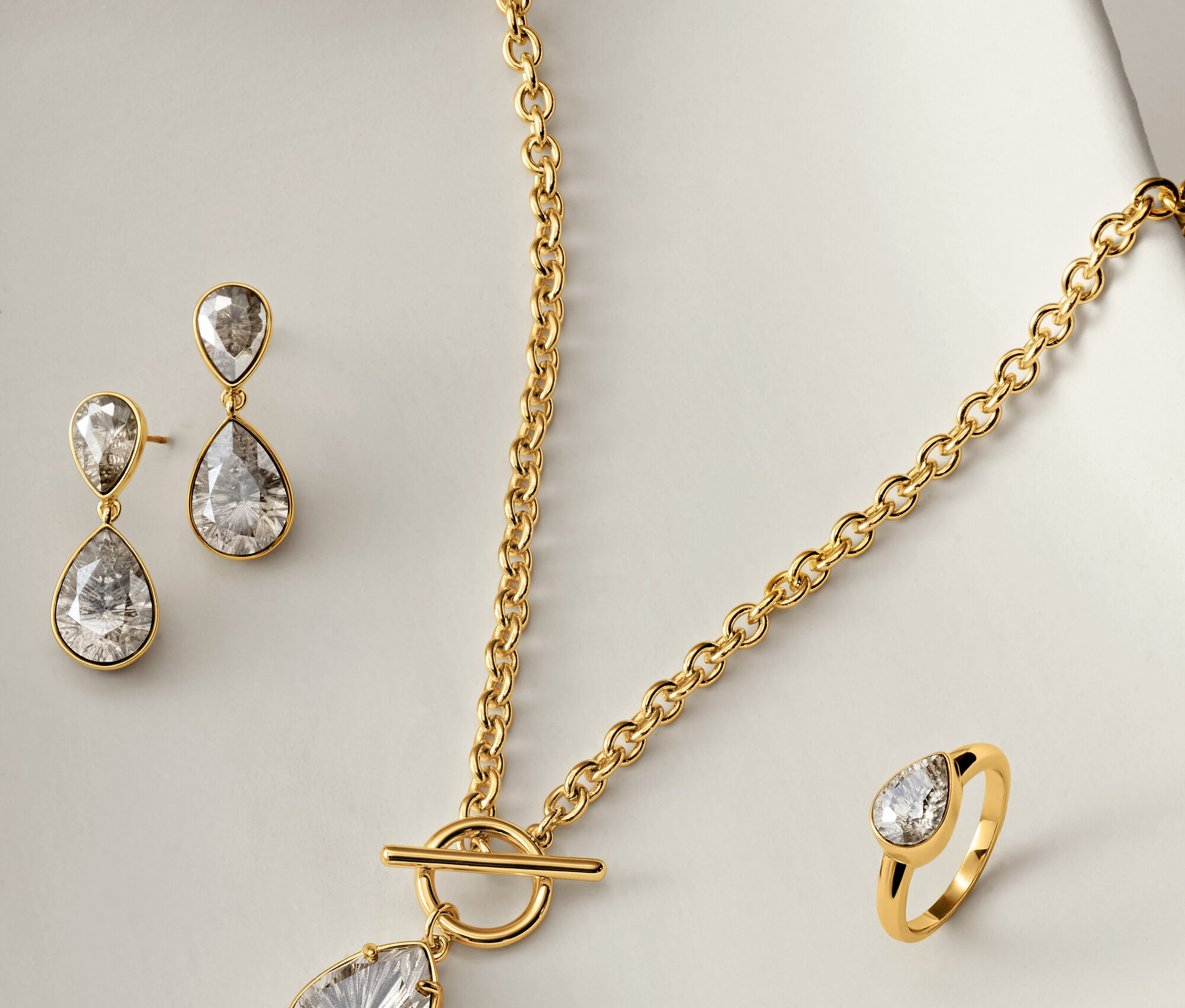 Goldene Halskette, Ohrringe und Ring mit klaren, tropfenförmigen Steinen auf weißem Hintergrund.