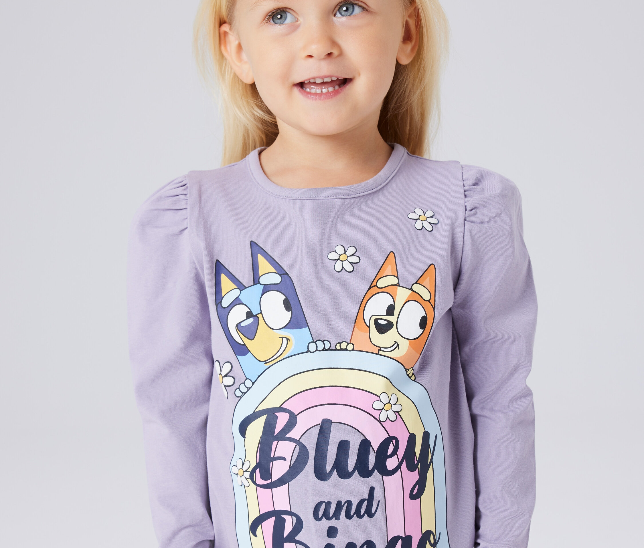 Ein kleines blondes Mädchen trägt ein langärmliges Shirt mit einem Aufdruck von Bluey und Bingo auf einem Regenbogen.
