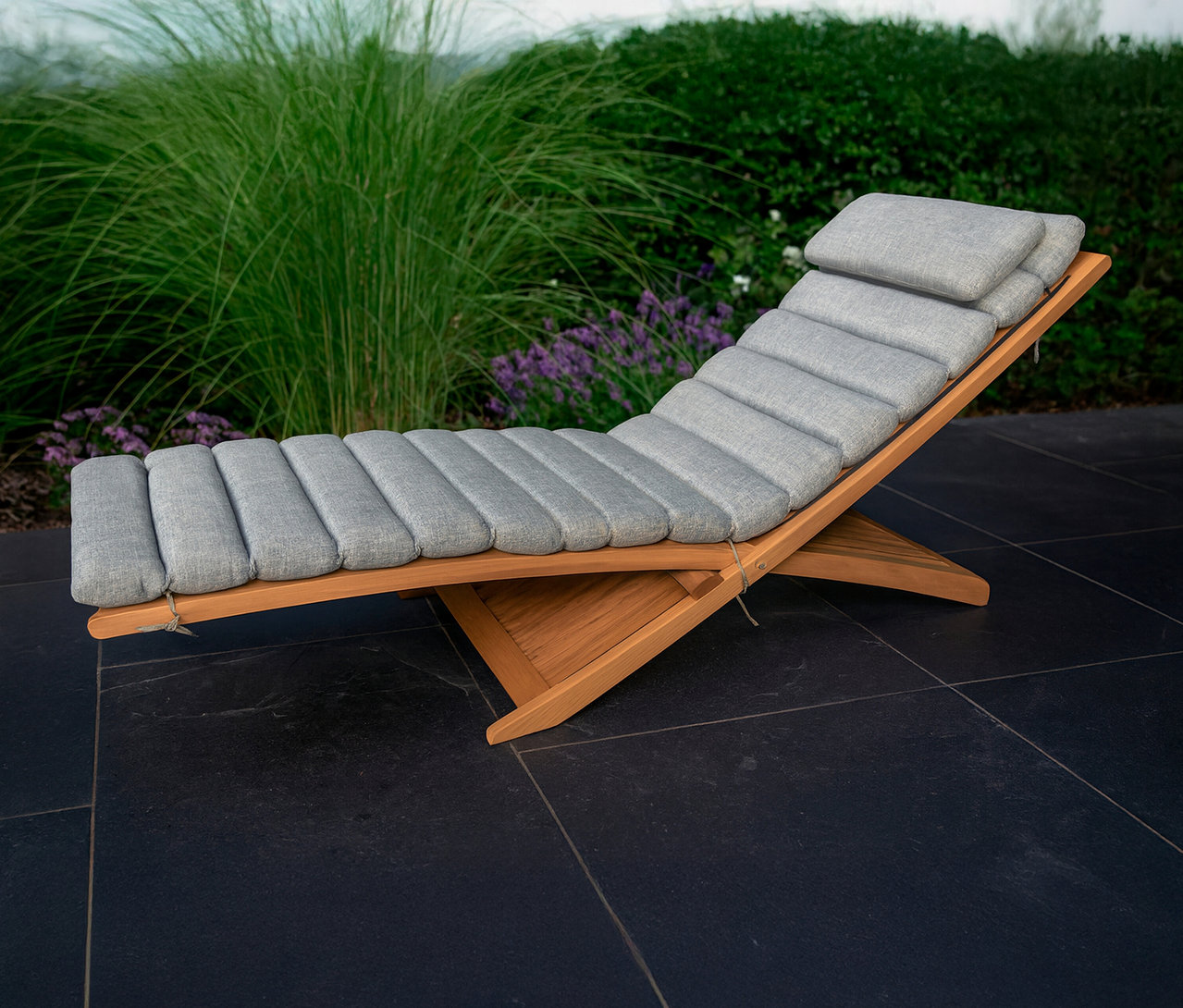 Eine Leco Sonnenliege mit grauen Polstern steht auf einer dunklen Terrasse vor Büschen.