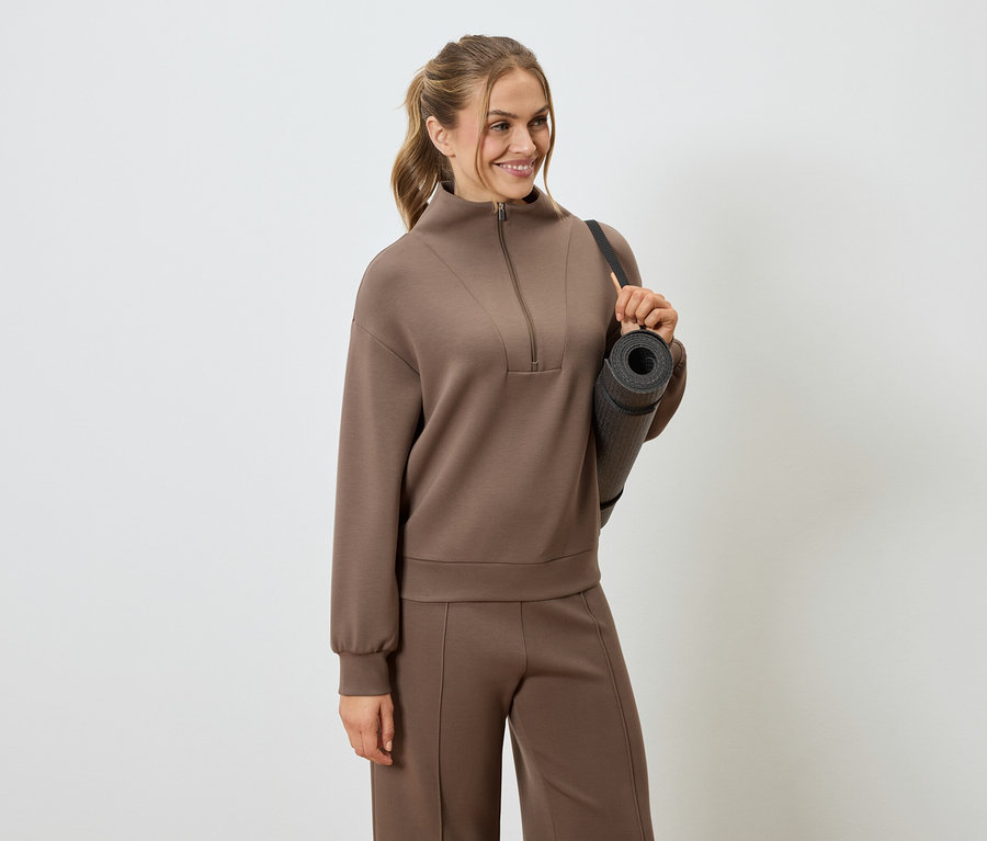 Frau trägt braunen Trainingsanzug und Yogamatte über der Schulter.