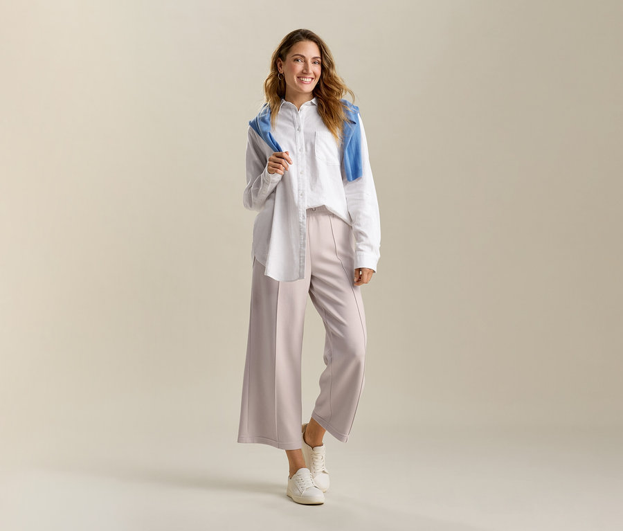 Frau mit weisser, langer Leinenbluse und sandfarbener Culotte aus Modal-Mix. Ein blauer Pullover liegt über ihren Schultern.