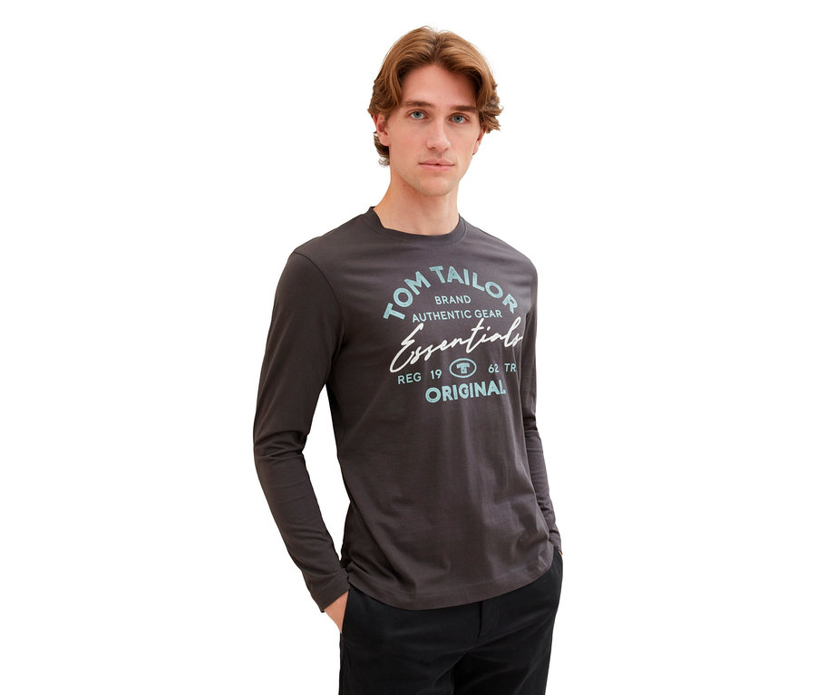 Mann mit braunen Haaren trägt ein graues Tom Tailor Langarmshirt und eine schwarze Hose.