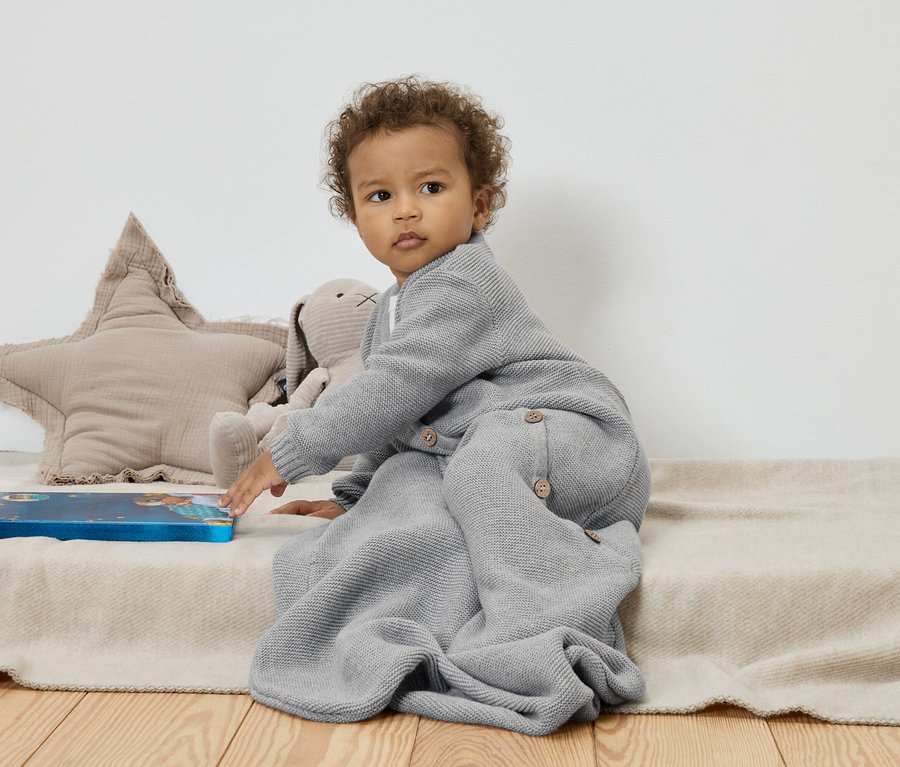 Baby sitzt auf einer beige Decke neben einem Stern- und Hasenkuscheltier und berührt ein blaues Buch.