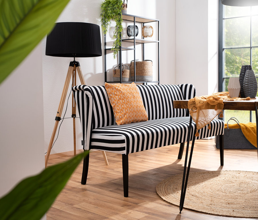 Innenaufnahme mit der schwarz-weiß gestreiften Polsterbank »Hendrick« mit orangefarbenem Kissen und Stehlampe. Im Hintergrund Regal und Tisch.