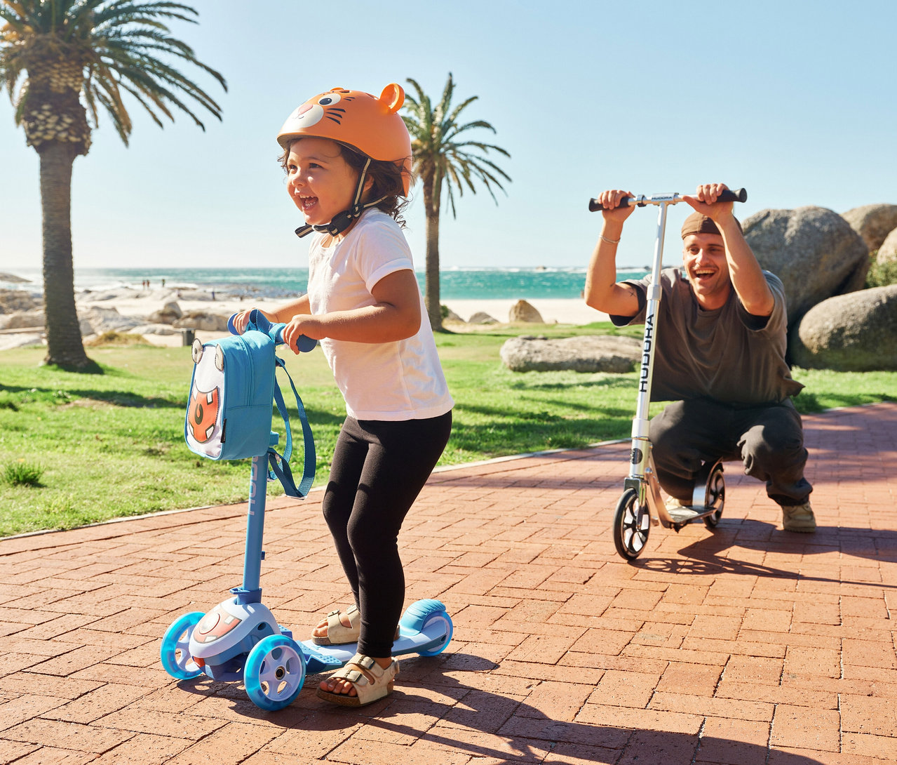 Kind mit Helm fährt Hudora 4-in-1-Scooter in Cyan, während ein Mann auf einem Roller daneben hockt. Palmen und Meer im Hintergrund.