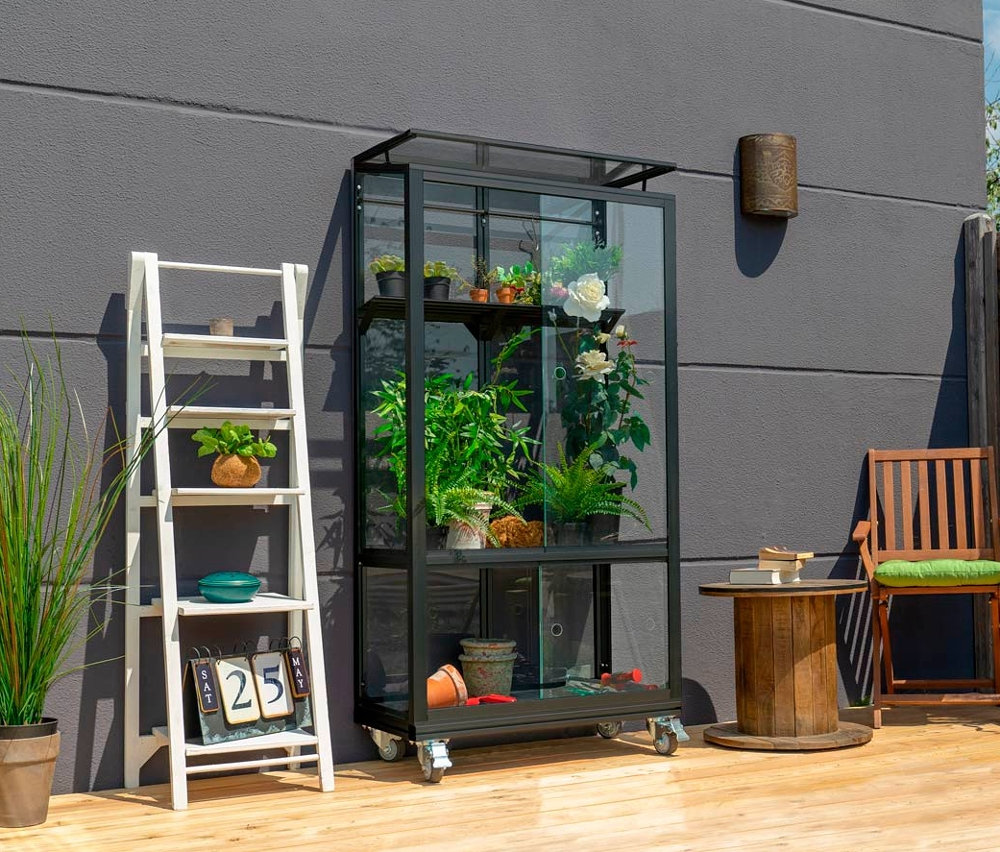 Ein schwarzes Vitavia Balkongewächshaus mit Rollfundament steht auf einer Holzterrasse neben einer weißen Leiter mit Pflanzen.