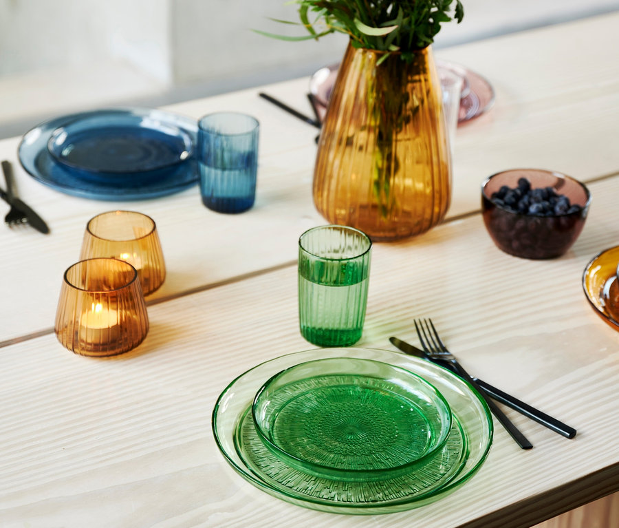 Auf einem Tisch sind grüne und blaue Kusintha-Glasteller von Bitz (25 cm), Gläser, eine Blumenvase und eine Schale mit Obst angerichtet.