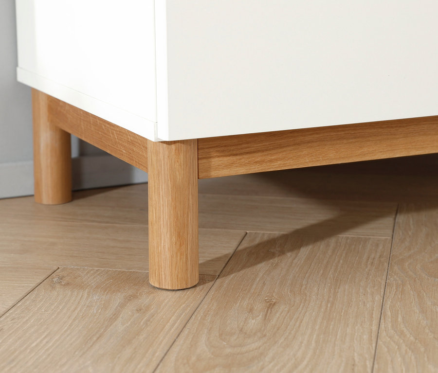 Detailaufnahme vom Sockel einer weißen Rauch Kommode »Freyja« mit Holzfüßen auf einem Holzboden.