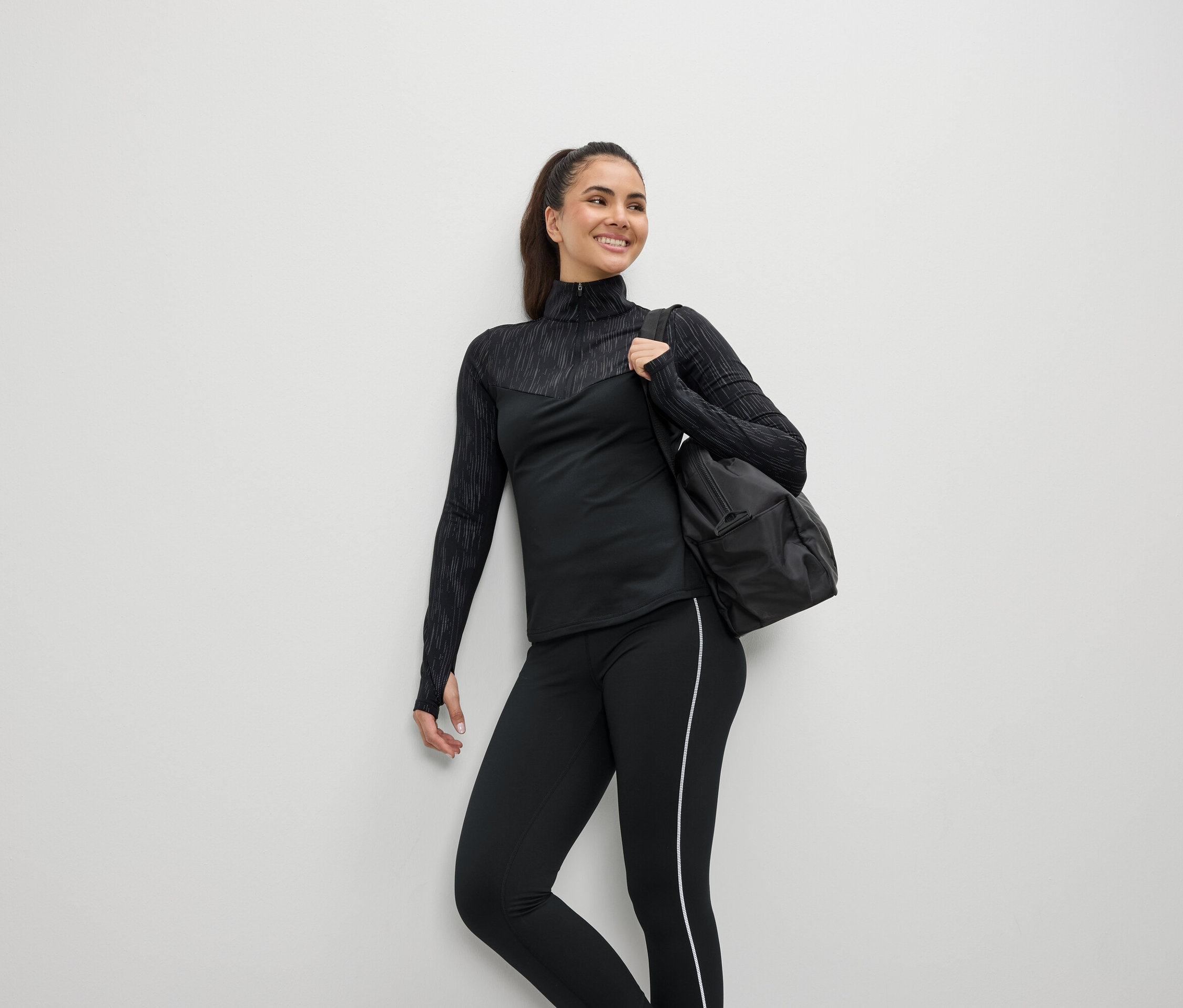Lächelnde Frau in schwarzer Sportbekleidung mit einer Tasche über der Schulter.