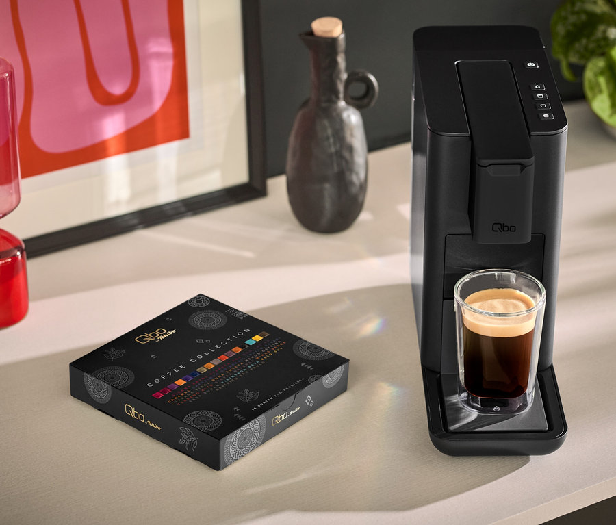 Eine Tasse Kaffee steht neben einer schwarzen Qbo ICONIC Dark Chrome und einer Qbo Collection - 16 Kapseln Packung.