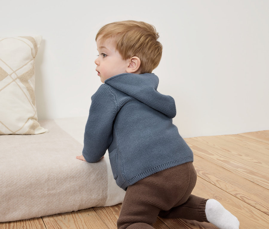 Baby krabbelt auf einer beigen Couch in einem blauen Kapuzenpullover und brauner Hose.