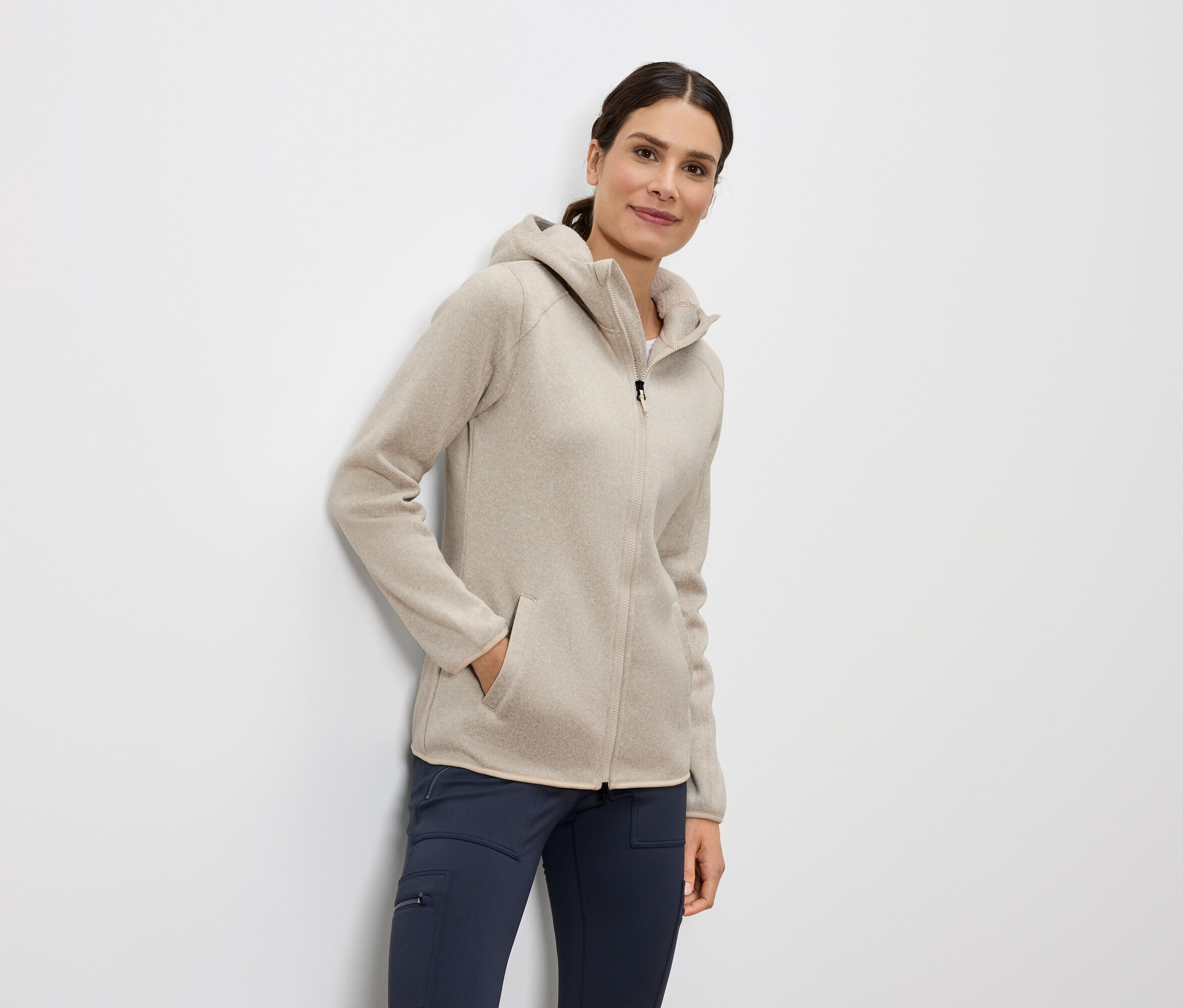 Frau steht mit der Hand in der Tasche einer beige Fleecejacke mit Kapuze und Reißverschluss und blauen Hosen.