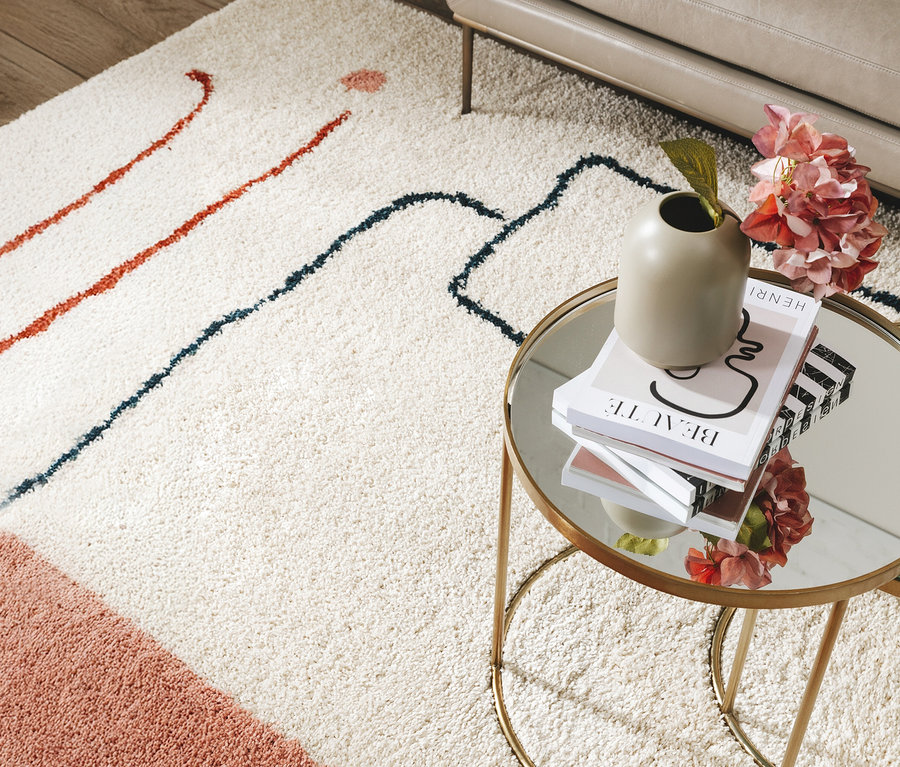 Ein beiger Teppich mit roten und blauen Linien liegt unter einem goldenen Beistelltisch mit Büchern, einer Vase und rosa Blumen.