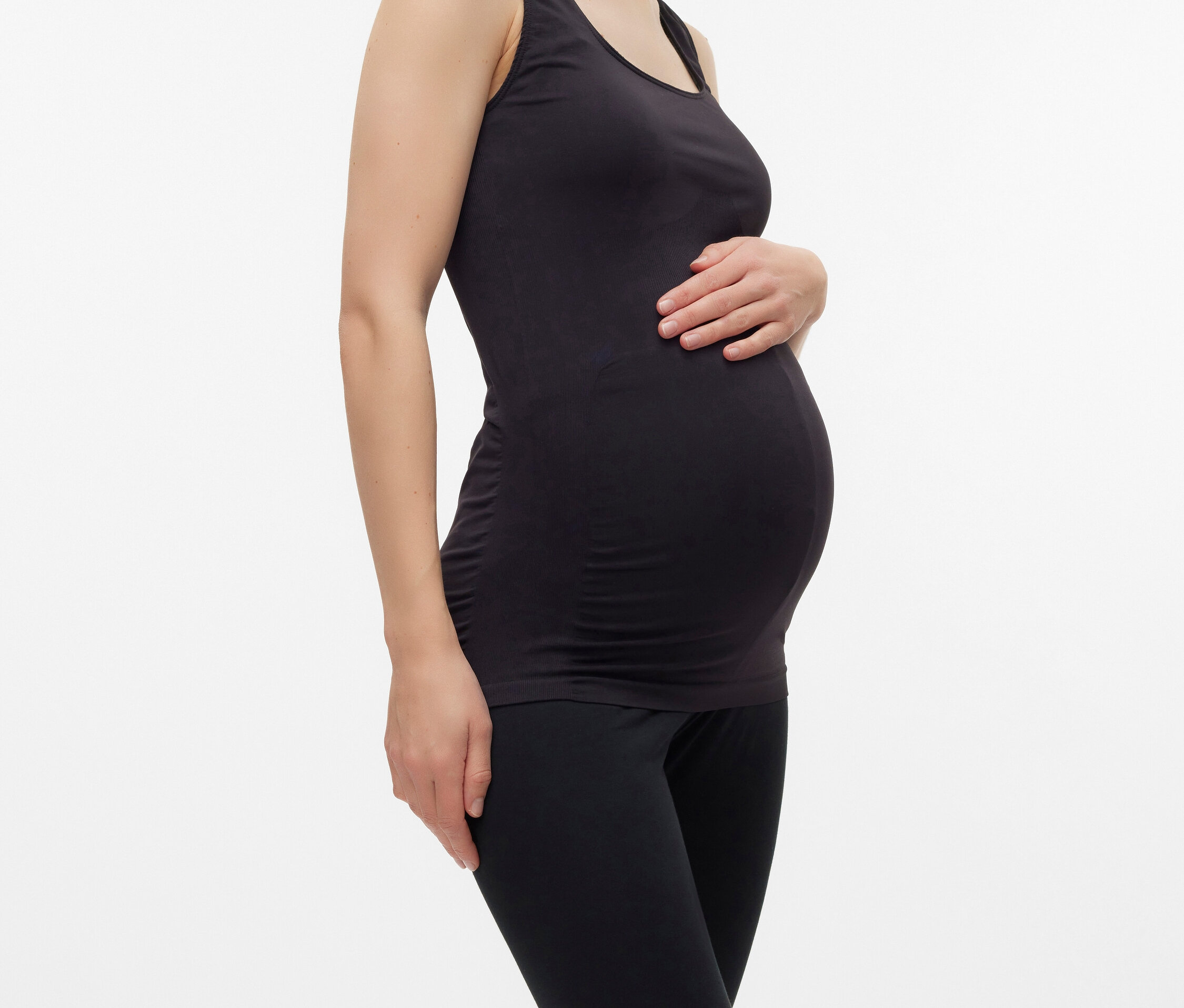 Schwangere Frau mit schwarzem Tanktop und Leggings posiert vor einem weißen Hintergrund.