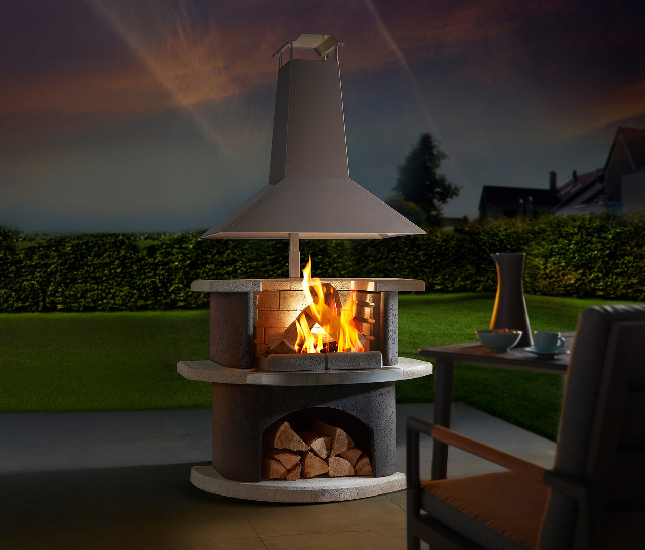 Feuer brennt in einem gemauerten Außenkamin mit Ablagefläche für Holz, im Hintergrund Gartenmöbel und eine grüne Hecke.