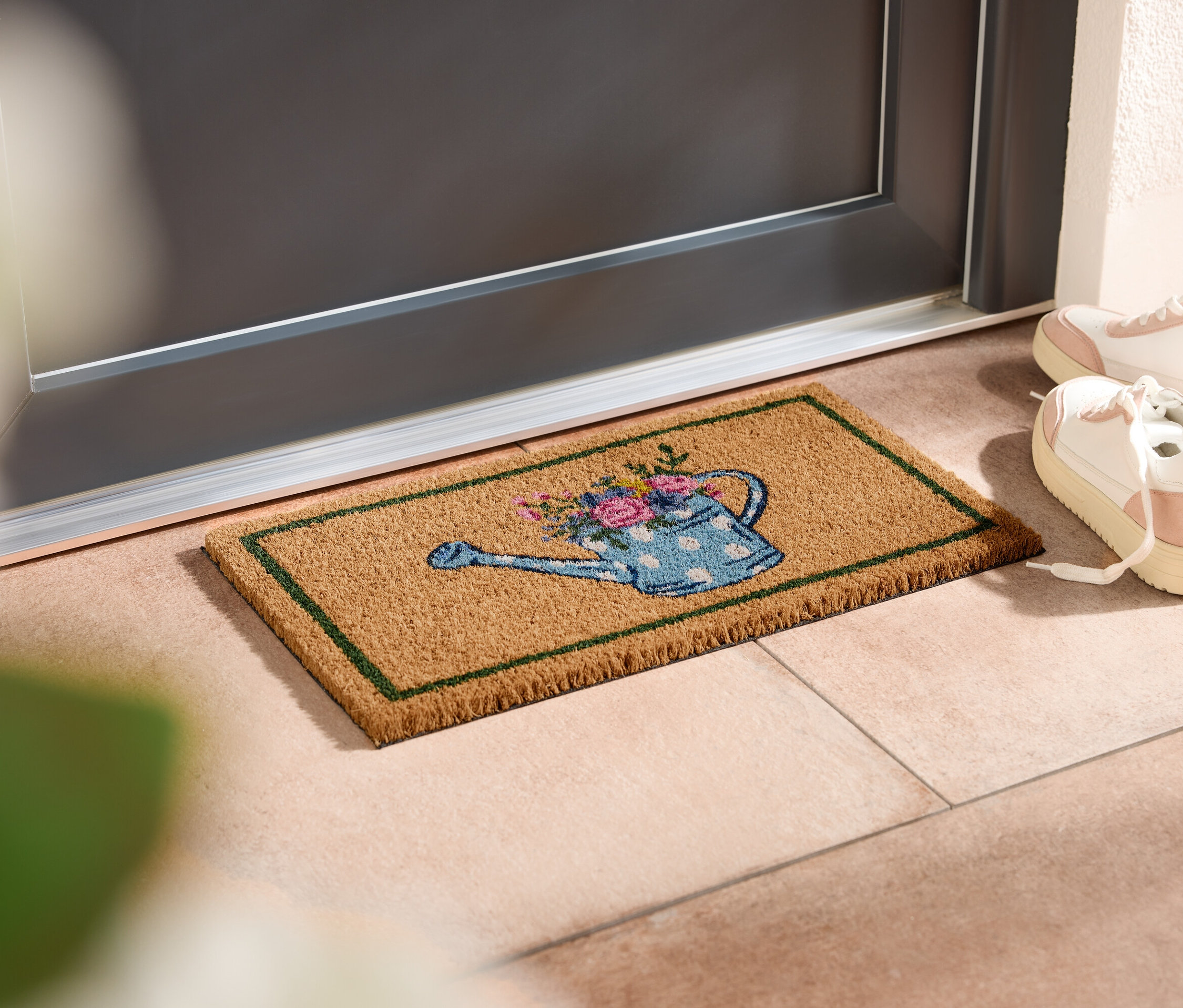 Eine Fußmatte mit Gießkannenmotiv liegt vor einer Tür, daneben stehen weiße Turnschuhe.