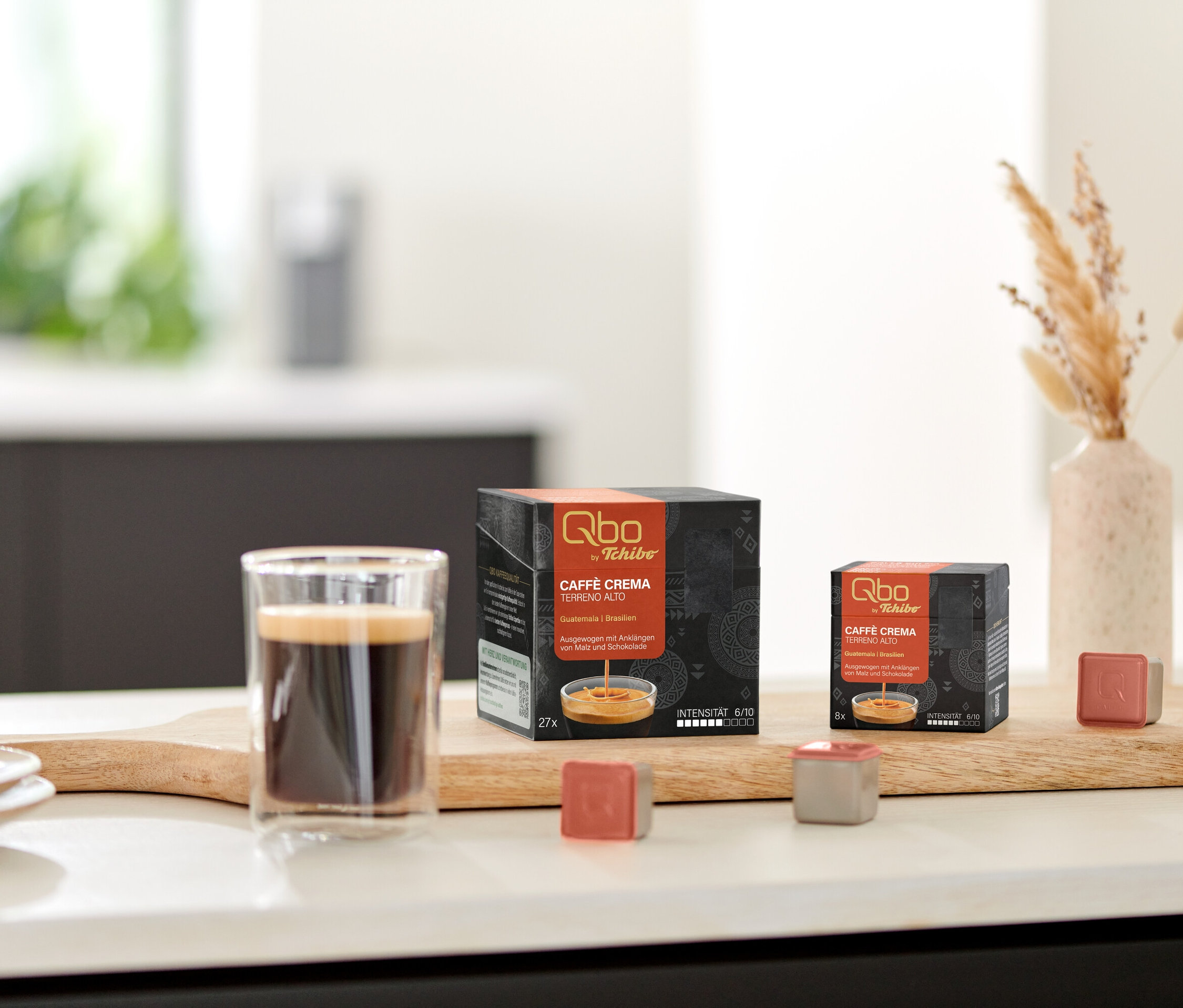 Auf einem Tisch stehen Qbo Caffè TERRENO ALTO Kapseln neben einem Glas Kaffee und Keksen.