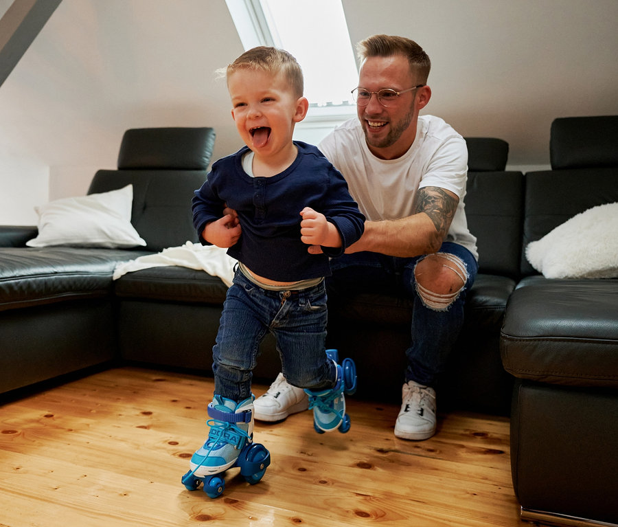 Ein Junge fährt auf cyanfarbenen HUDORA Rollschuhen »My First Quad« und lacht, während ein Erwachsener auf ihn aufpasst.