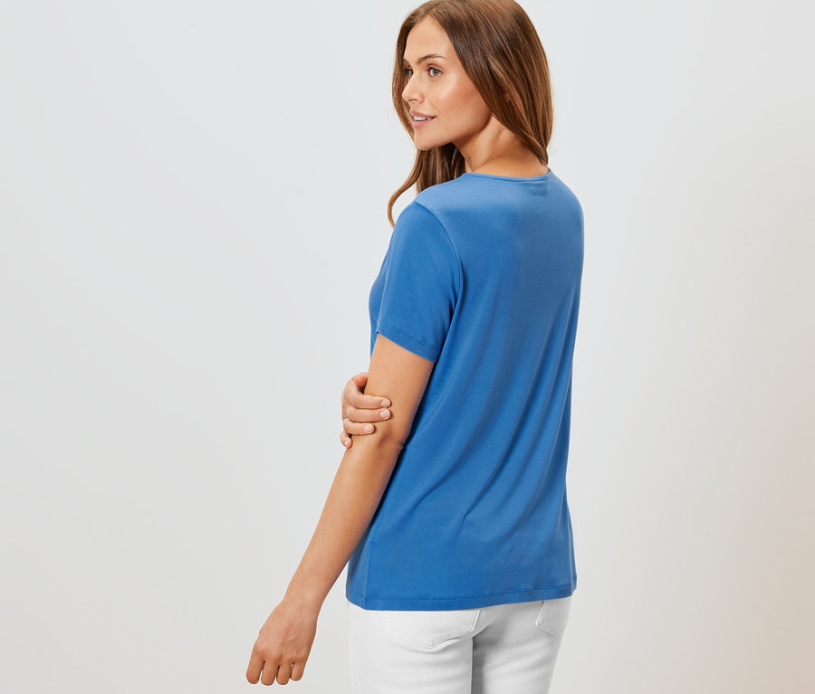 Frau posiert in kobaltblauem Shirt mit Faltendetail und weißer Hose.