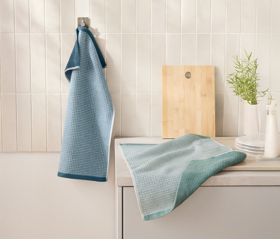 Ein blaues 2 Waffelpiqué-Küchenhandtücher hängt an einem Haken neben einem Schneidebrett. Ein weiteres Küchentuch liegt auf einer Küchentheke.