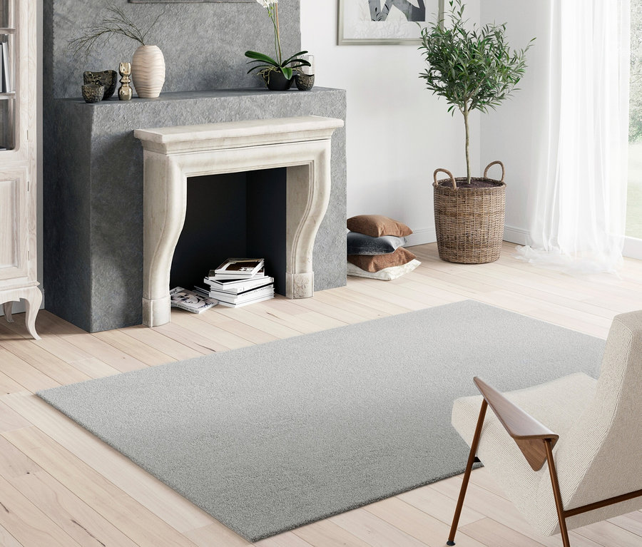 Ein hellgrauer Teppich »Mellow« liegt in einem Wohnzimmer mit Kamin und Sessel auf einem Holzboden.