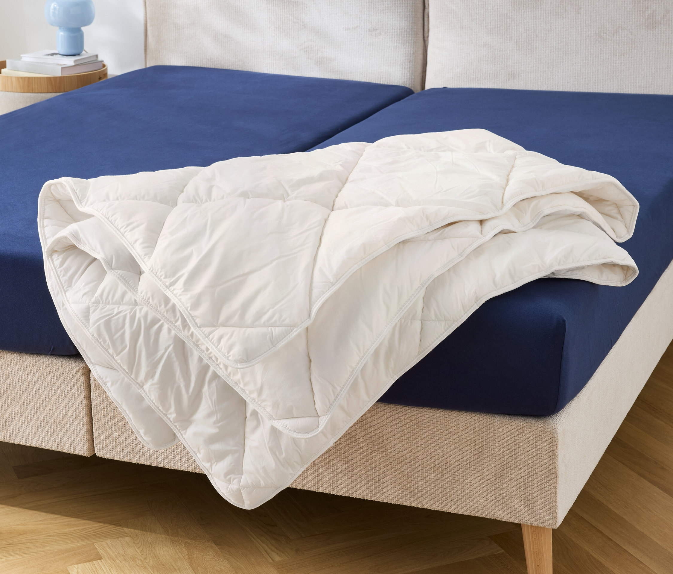 Auf einem Bett mit blauem Spannbettlaken liegt eine weiße Steppdecke.