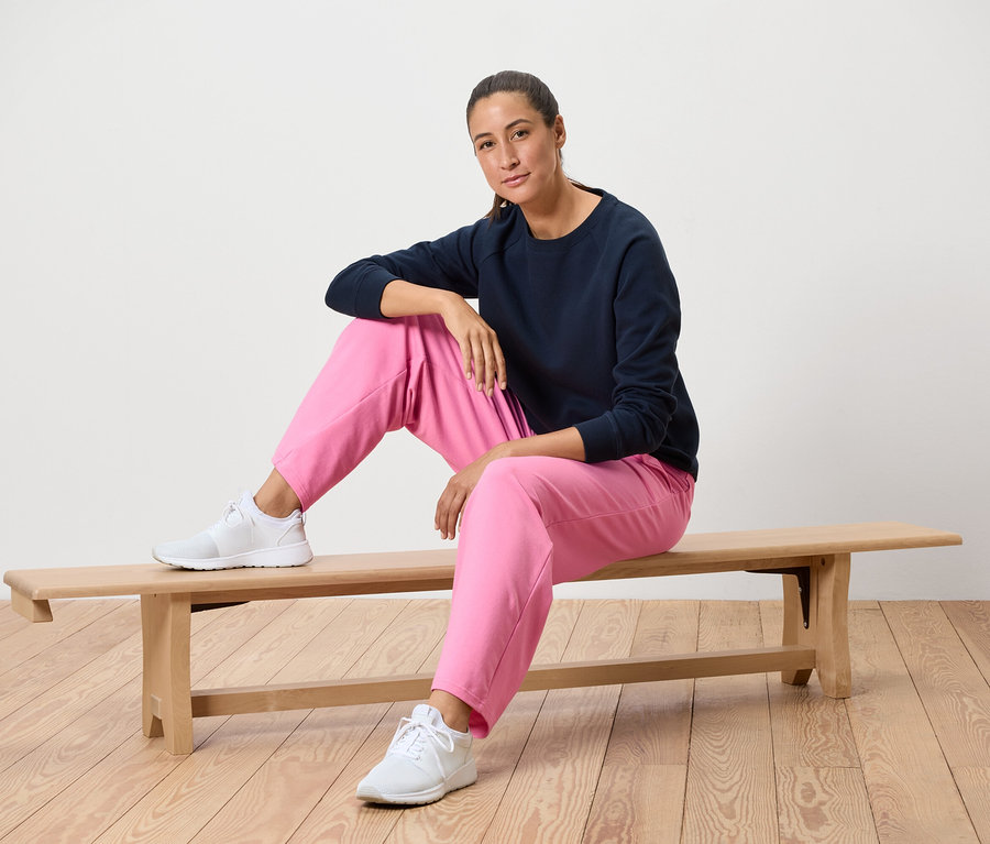 Frau sitzt auf einer Holzbank, trägt ein dunkelblaues Sweatshirt, eine pinke Hose und weiße Turnschuhe.