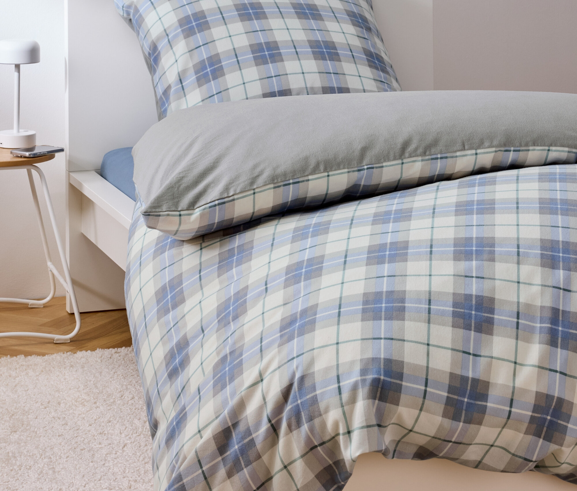 Bett mit blau-weiß karierter Bettwäsche und grauem Kissen.