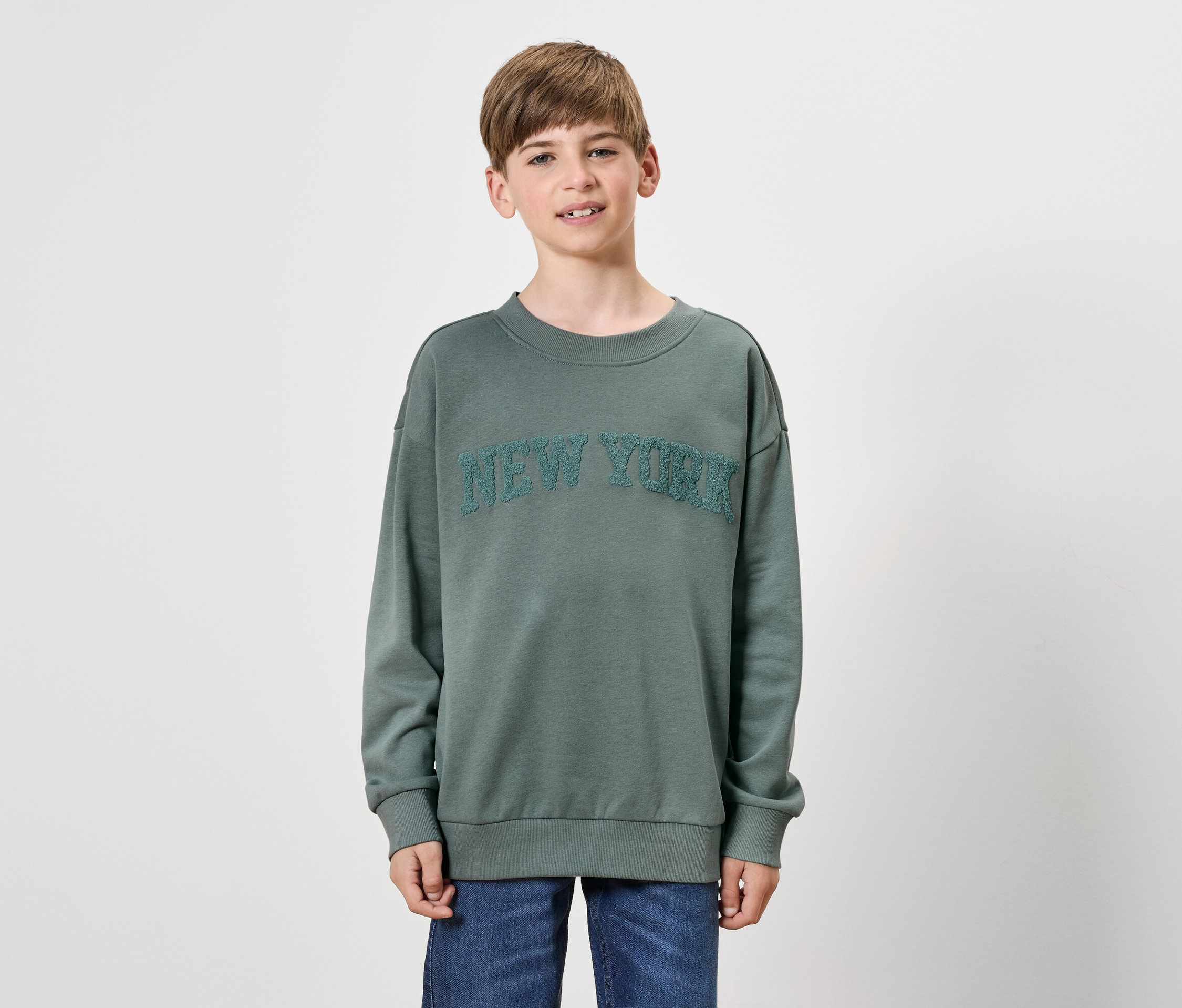 Ein Junge trägt einen grünen Pullover mit der Aufschrift NEW YORK und blaue Jeans.