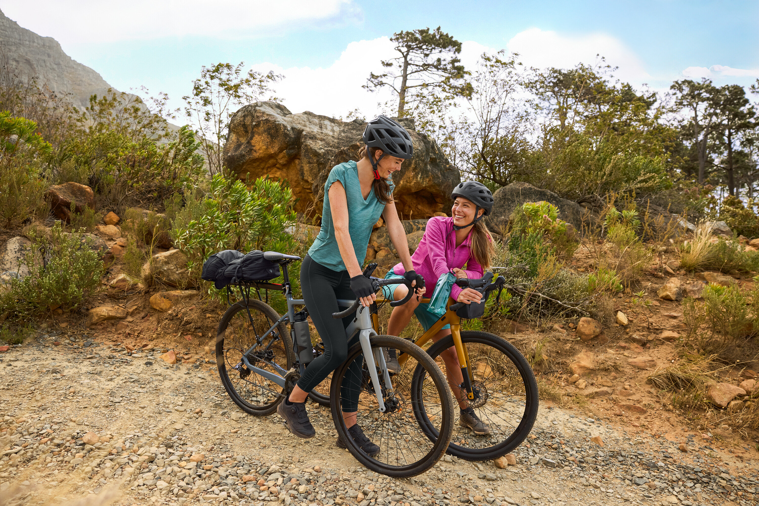 Zwei Frauen mit Helmen stehen mit ihren Fahrrädern auf einem Schotterweg in hügeligem Gelände. Eine Trinkflasche ist am Fahrrad befestigt.