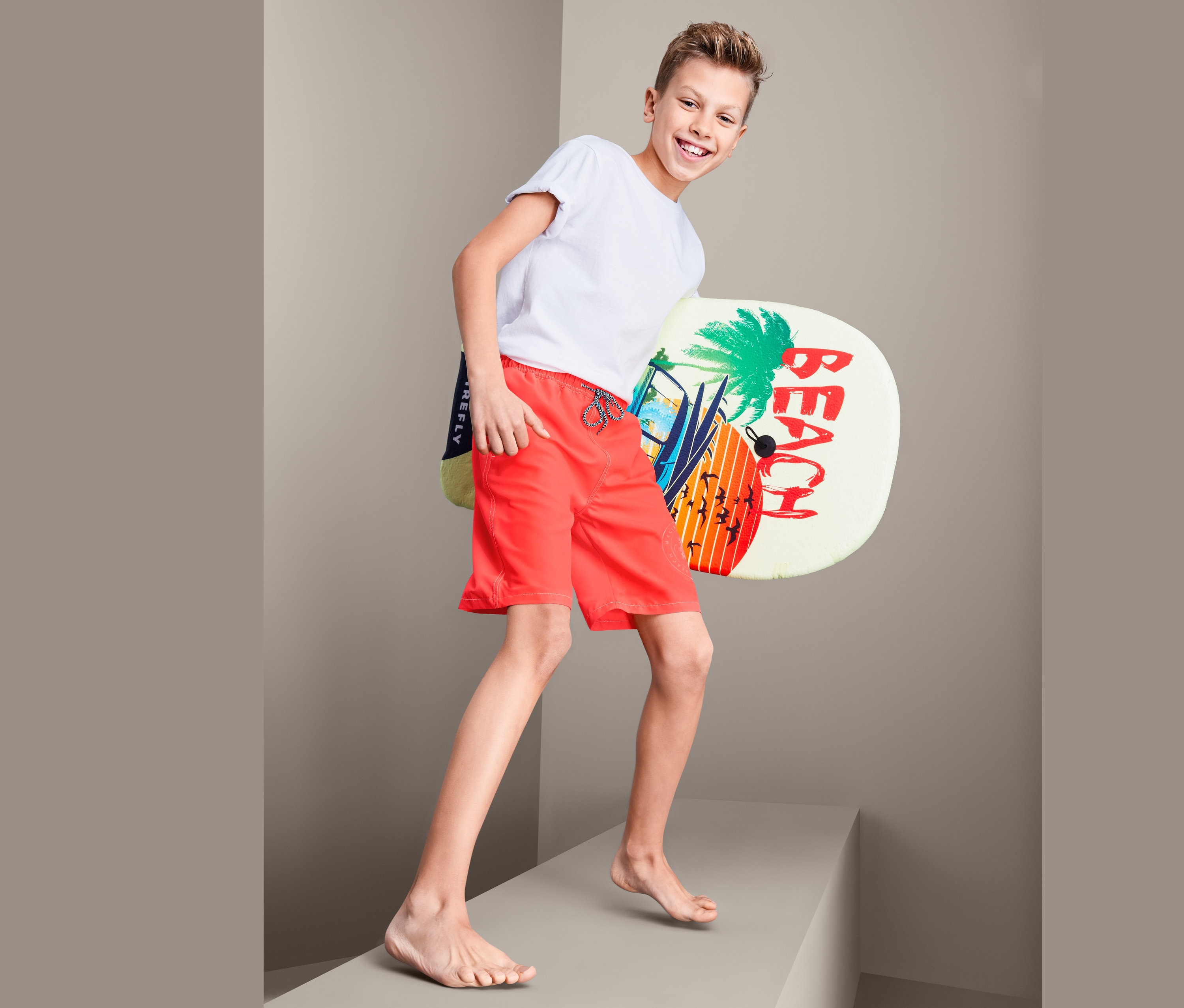 Ein Junge in roten Boardshorts hält ein Brett mit Strandmotiv.