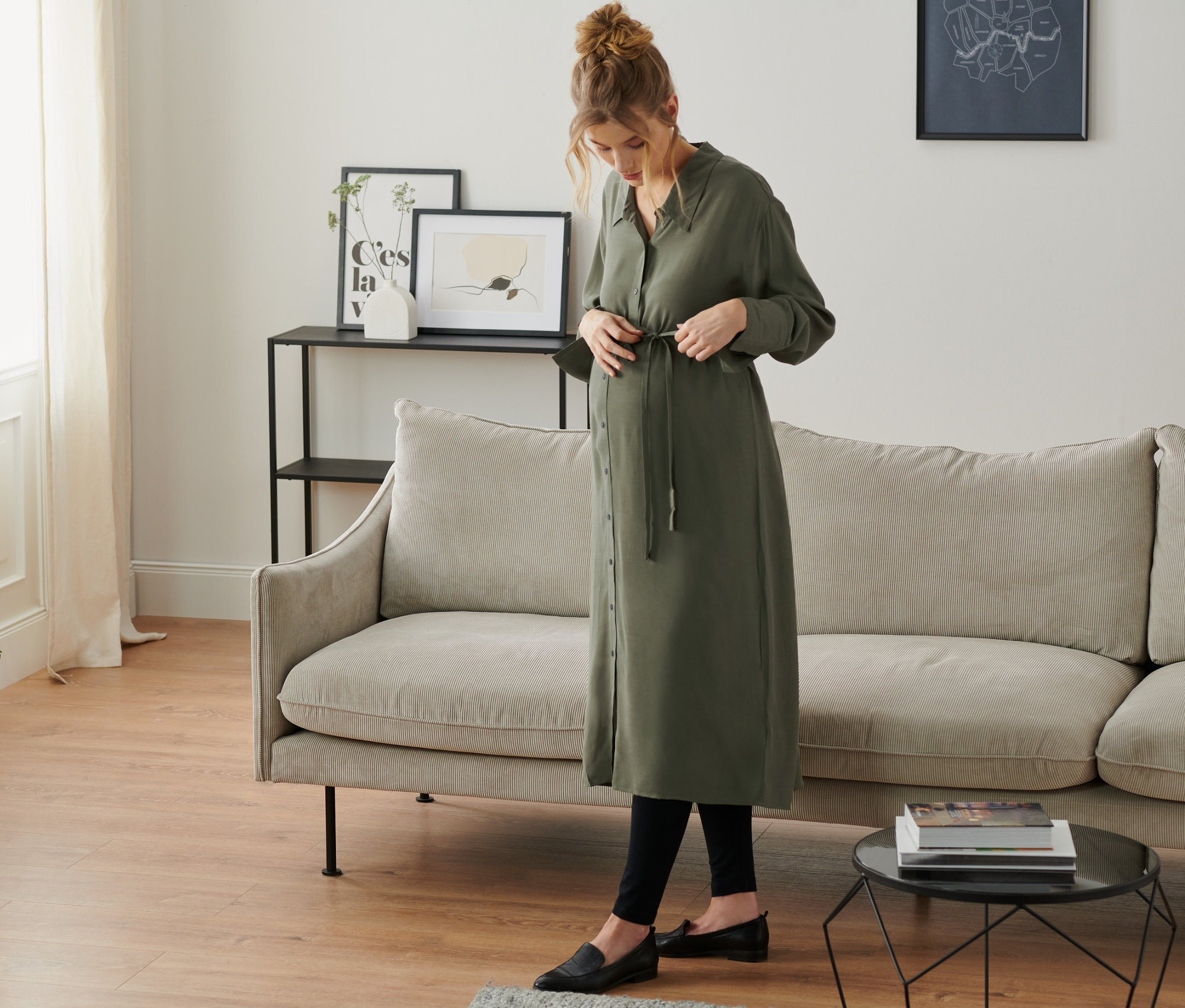 Eine schwangere Frau bindet ein olivgrünes Umstands-Blusenkleid in einem Wohnzimmer.