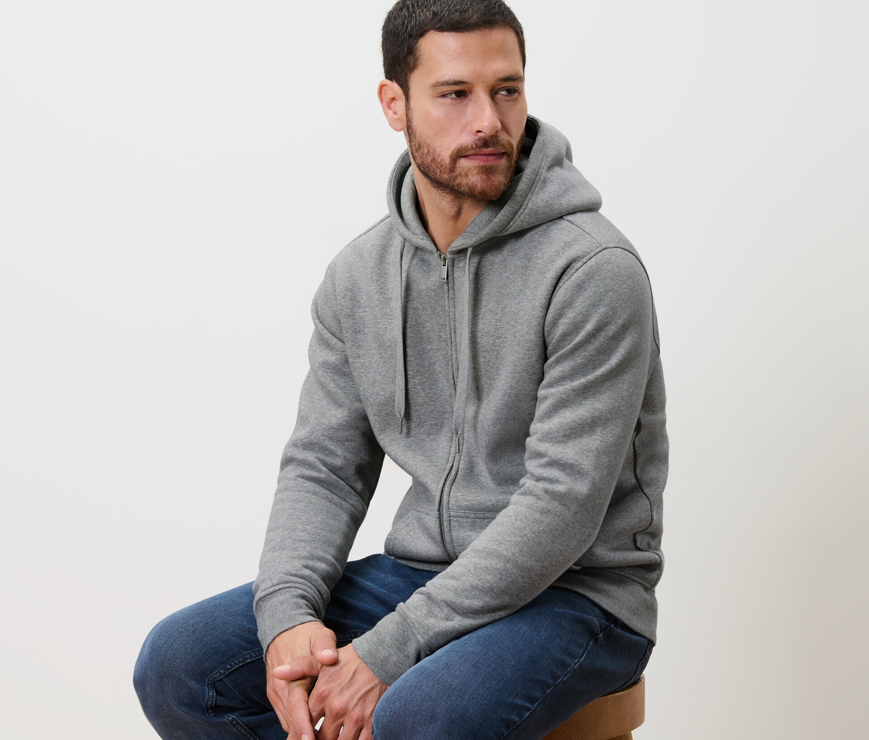 Mann sitzt auf einem Hocker und trägt eine graue Kapuzenjacke.