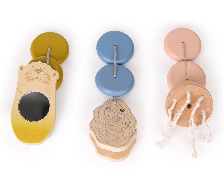 Drei Babyspielzeuge hängen nebeneinander. Eines ist gelb mit einem Otter und einem Spiegel, eines ist blau mit einer Holzzeichnung und eines ist rosa mit Schnüren.