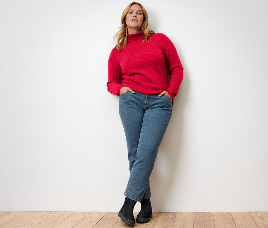 Frau posiert in geripptem, rotem Rollkragenpullover, Jeans und schwarzen Stiefeln.