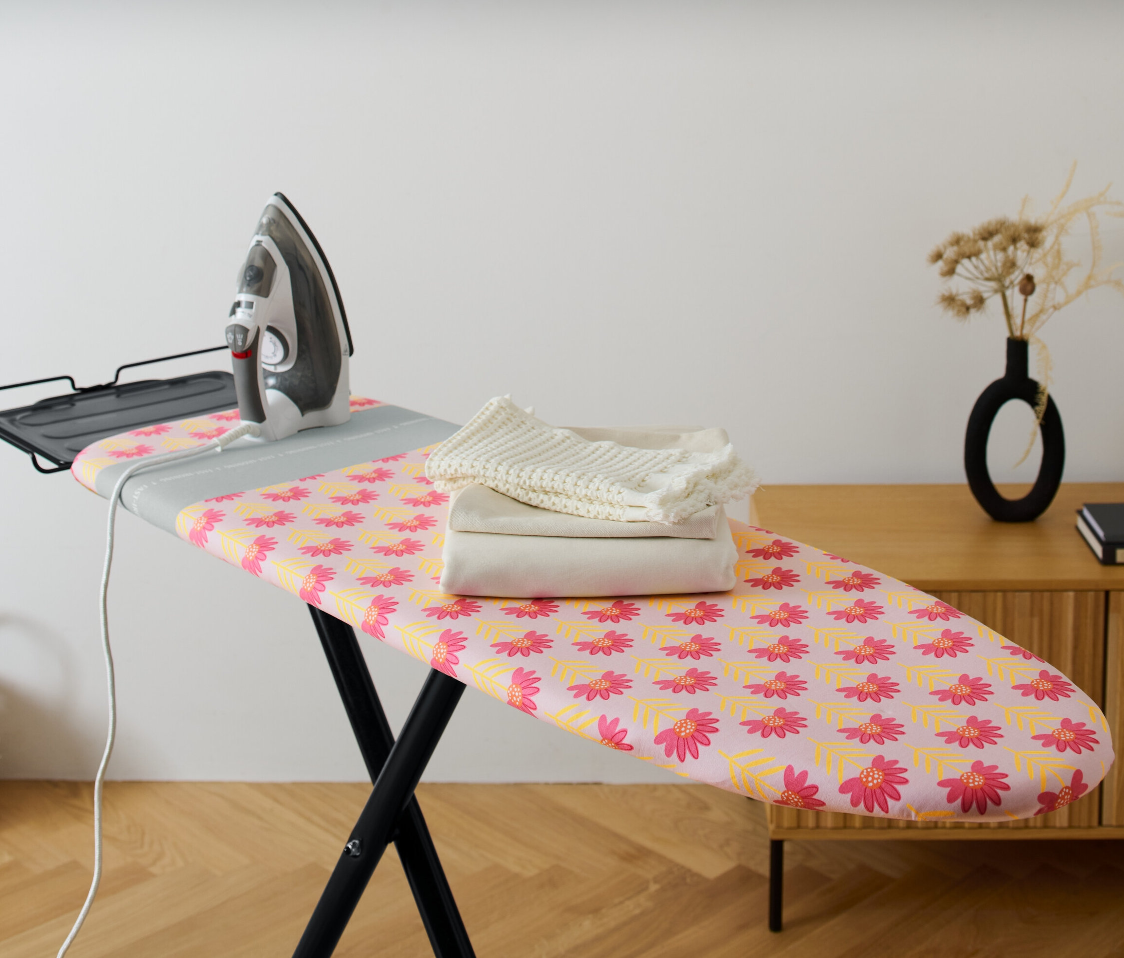 Ein Bügeleisen steht auf einem Bügelbrett mit einem blumigen Bügelbrettbezug. Auf dem Bügelbrett liegt gefaltete Wäsche.